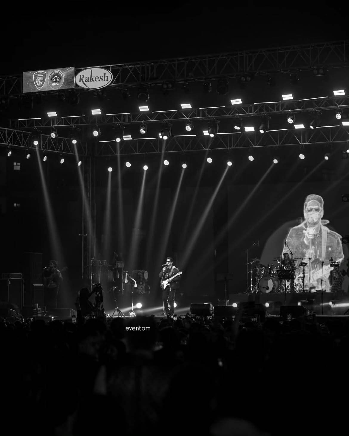 Black-and-white view of the Amit Mishra Live stage, beams falling through the truss onto the band and crowd.