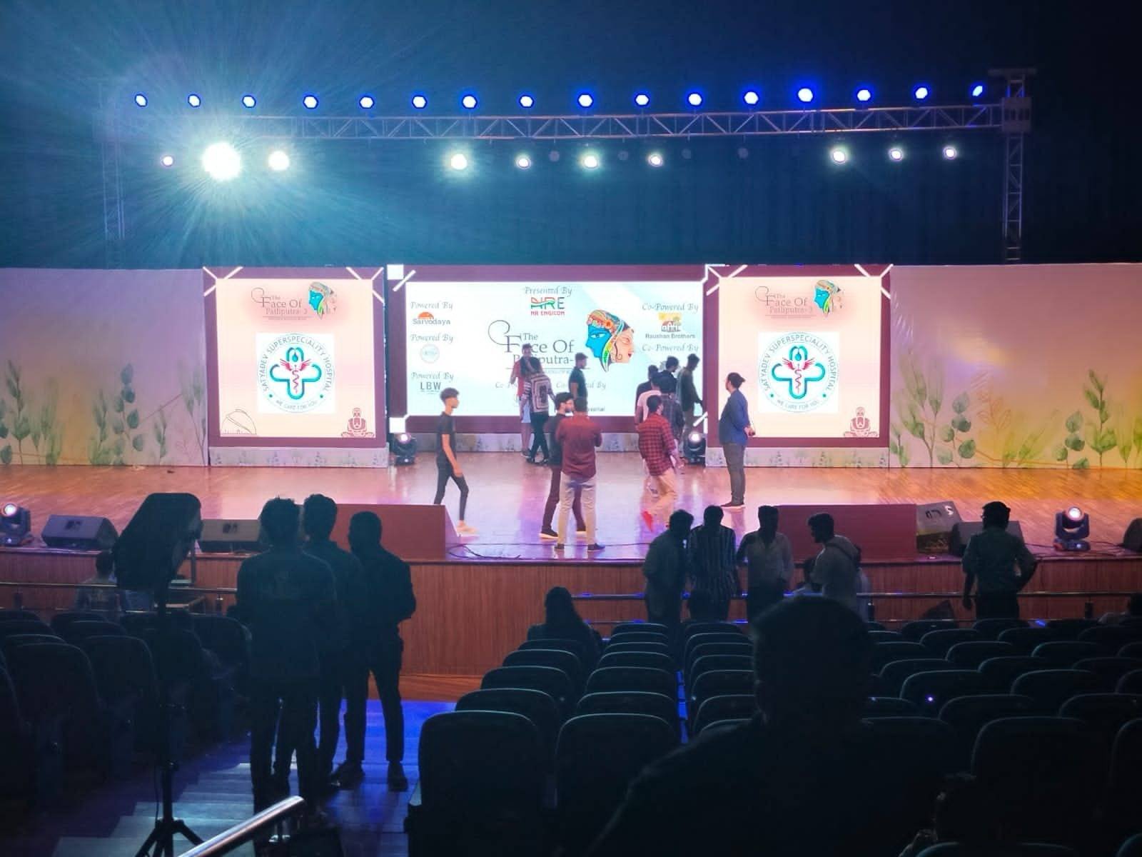 Bapu Sabhagar auditorium stage with a lighting truss, three branded LED screens and performers, seen from the seating.