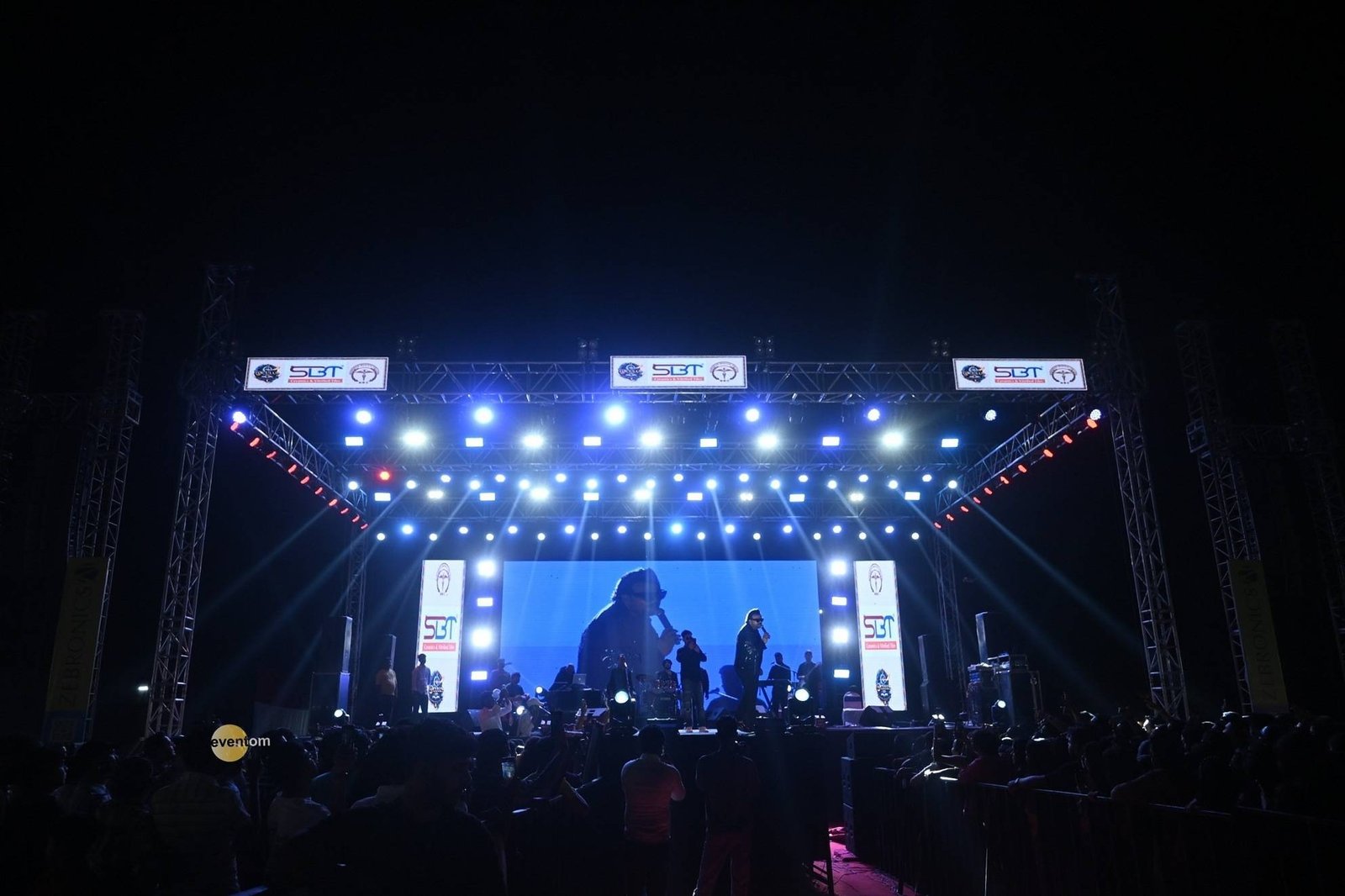 Outdoor concert stage washed in blue light, with truss towers, an LED wall and crowd silhouettes.
