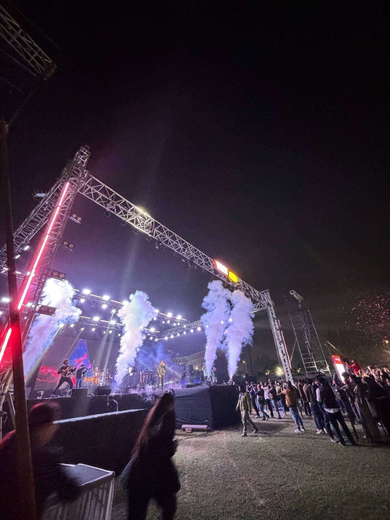 A large outdoor truss rig with CO₂ jets and stage lighting, seen from the edge of the crowd.