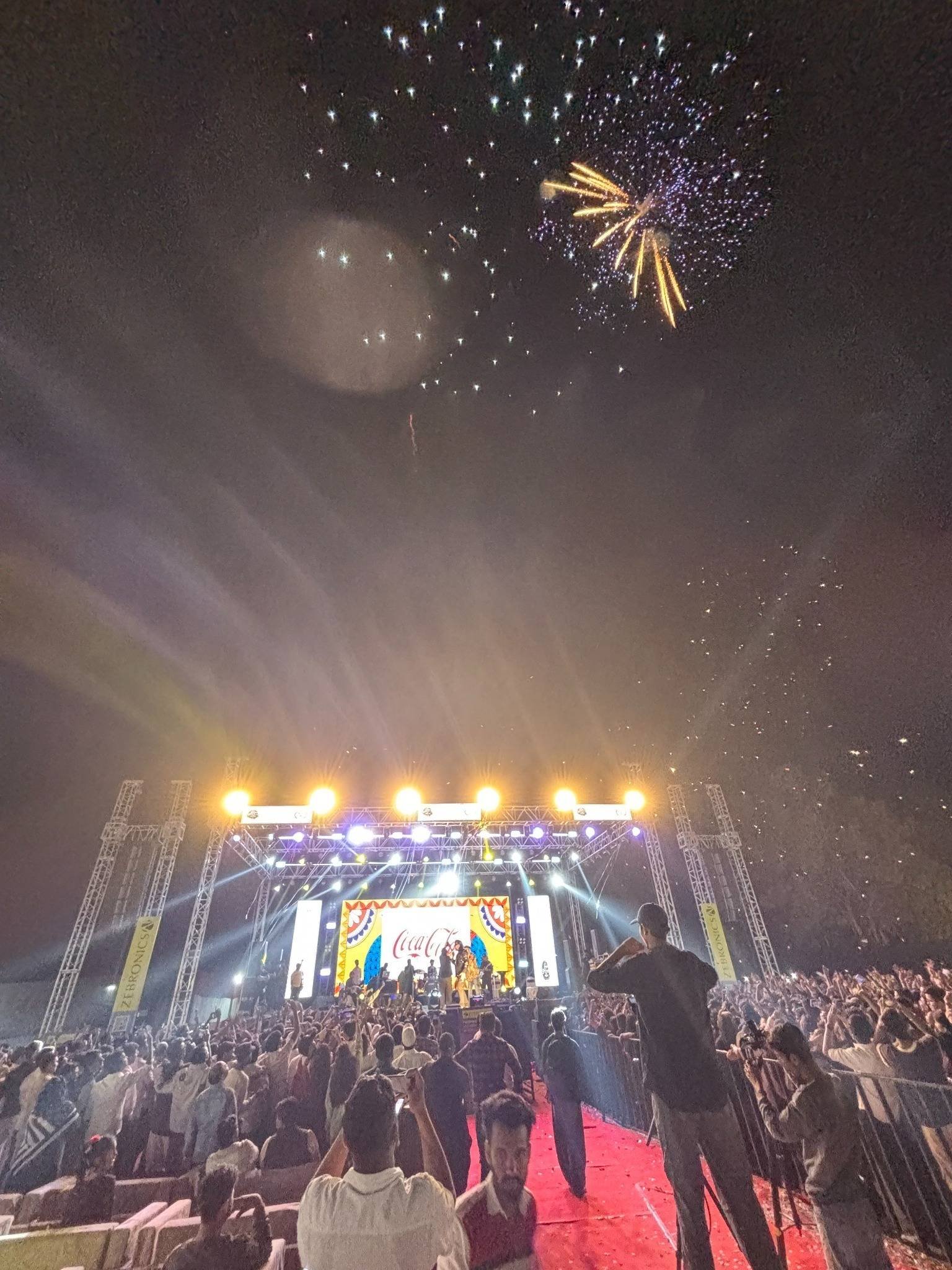 Fireworks burst above an outdoor concert stage as light beams cut through the haze over a packed crowd.