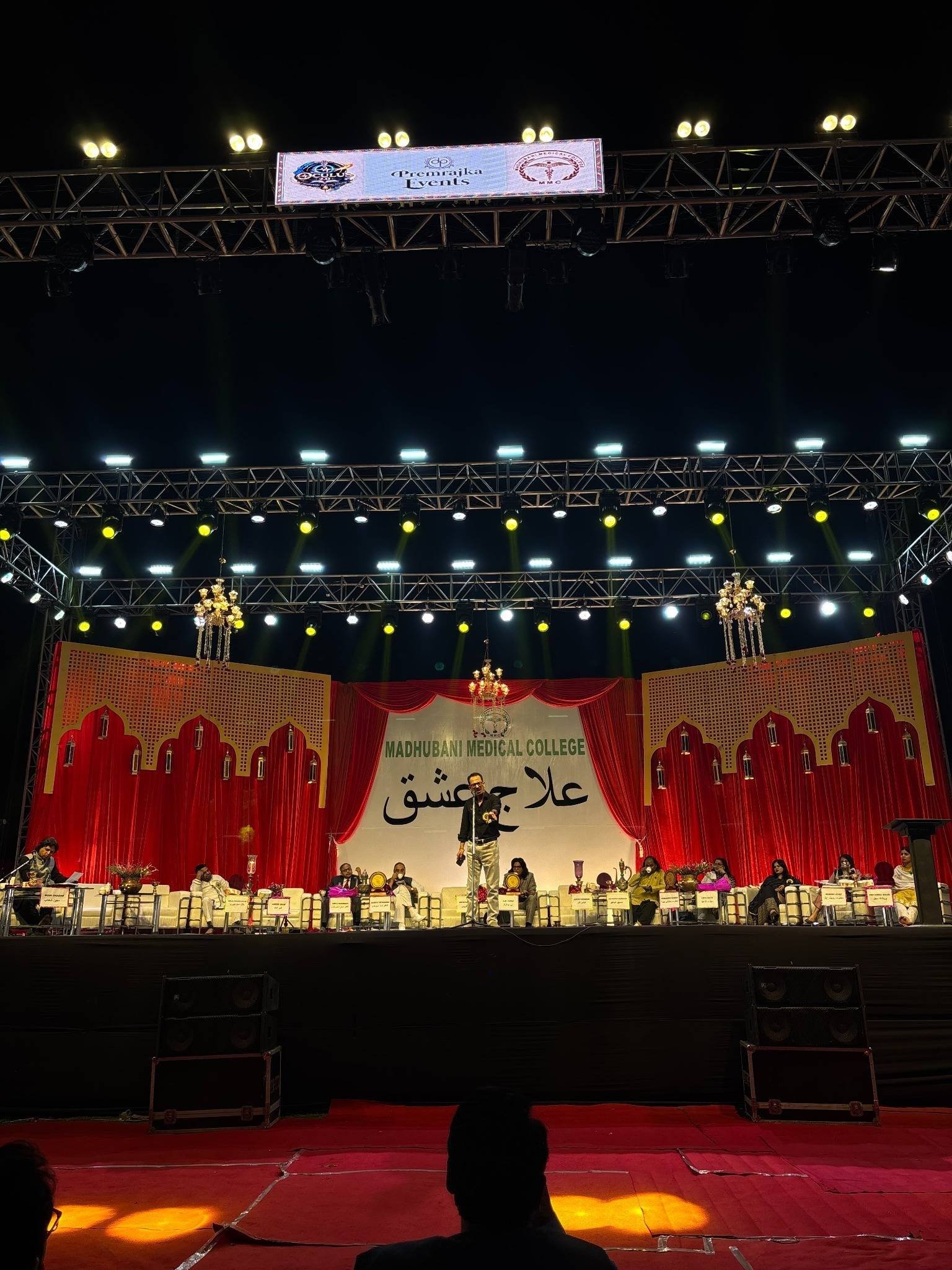 Madhubani Medical College stage under heavy lighting truss, with a red-and-gold arched backdrop and a Premrajka Events banner on the truss.