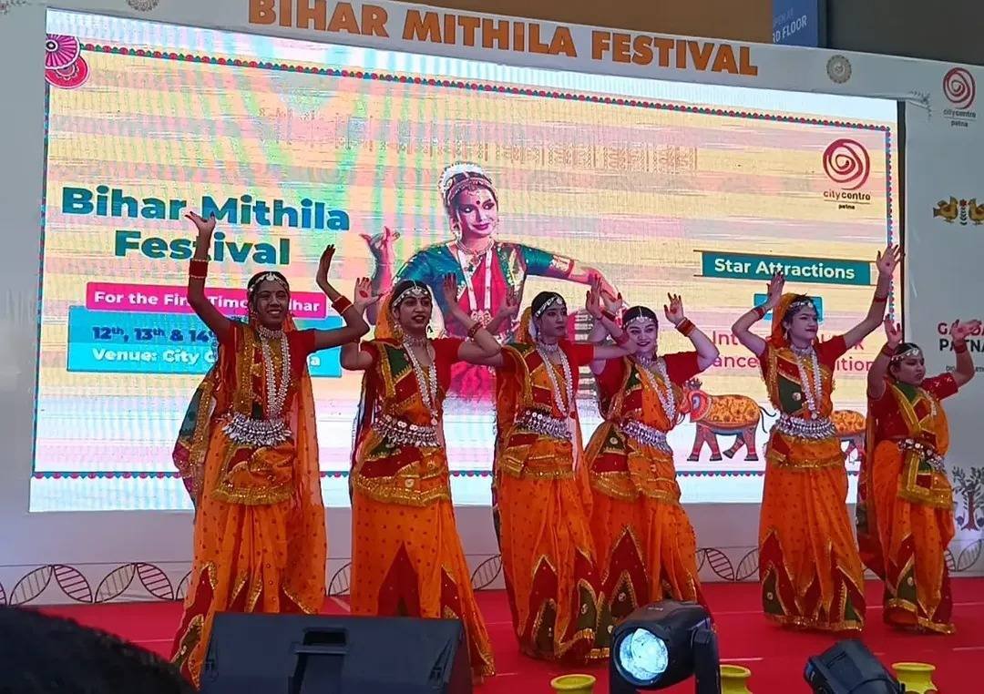 Dancers in orange costumes perform before the Bihar Mithila Festival LED backdrop.